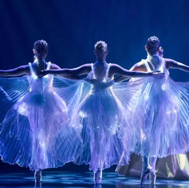hree ballet dancers on stage wearing illuminated flowing costumes under dramatic lighting.