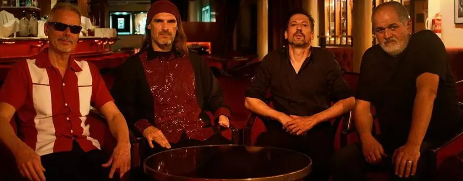 Four musicians seated around a small table in a warmly lit bar, posing for a group photo.