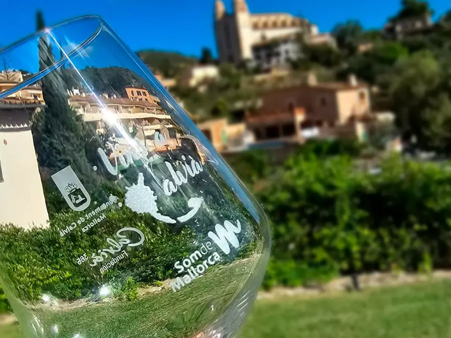 Wine glass with the logo of “Vins a Calvià” and Som de Mallorca, held up with the village of Calvià and its church in the background under a clear blue sky.