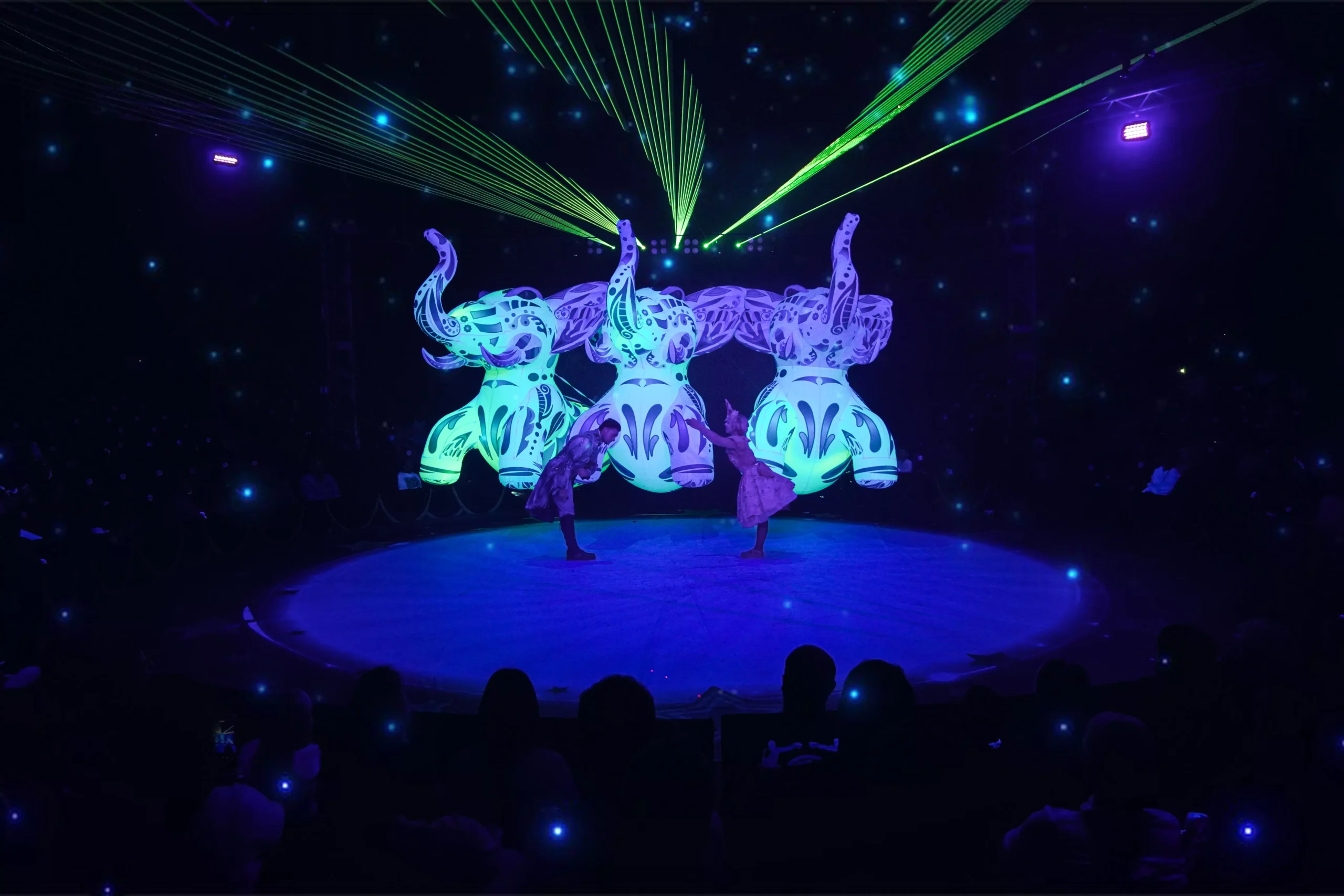 Three glowing elephant figures and acrobats performing under laser lights inside a dark circus tent.