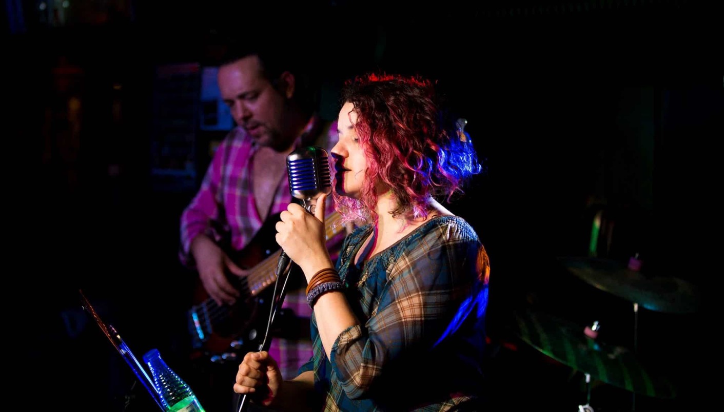 A singer performing with a vintage microphone on stage while a musician plays guitar behind her under colourful stage lighting.