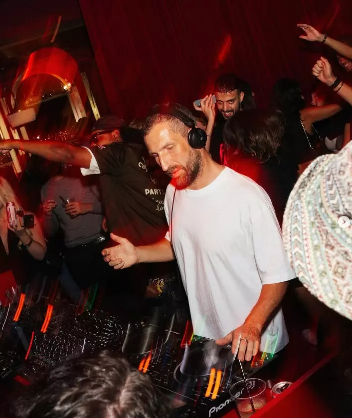 DJ Matthias Tanzmann performing in a red-lit nightclub, wearing headphones and a white T-shirt, surrounded by dancing clubgoers.