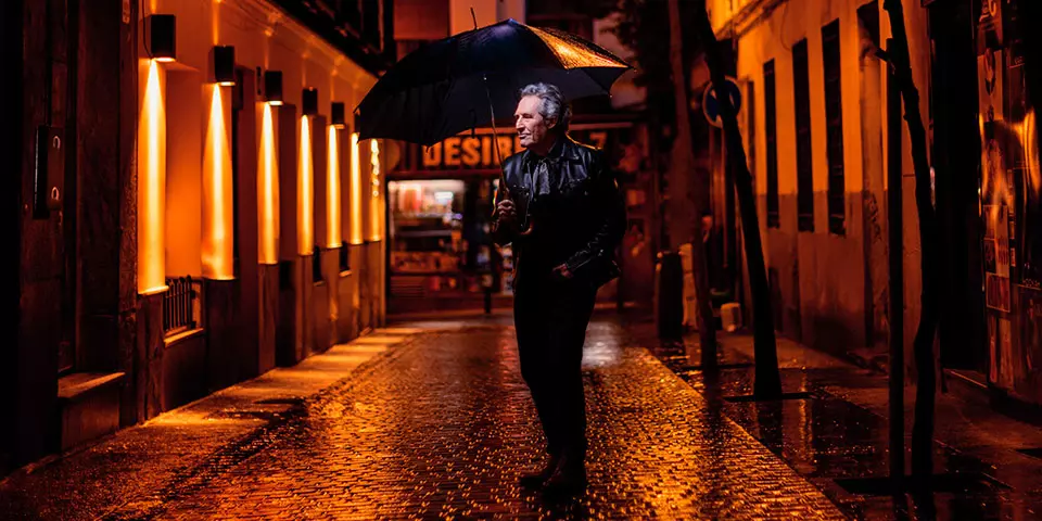 Miguel Ríos walking through a city street at night holding an umbrella
