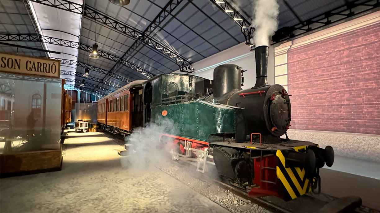 Indoor scene with a vintage steam locomotive emitting smoke, displayed on tracks inside a large covered railway museum hall.