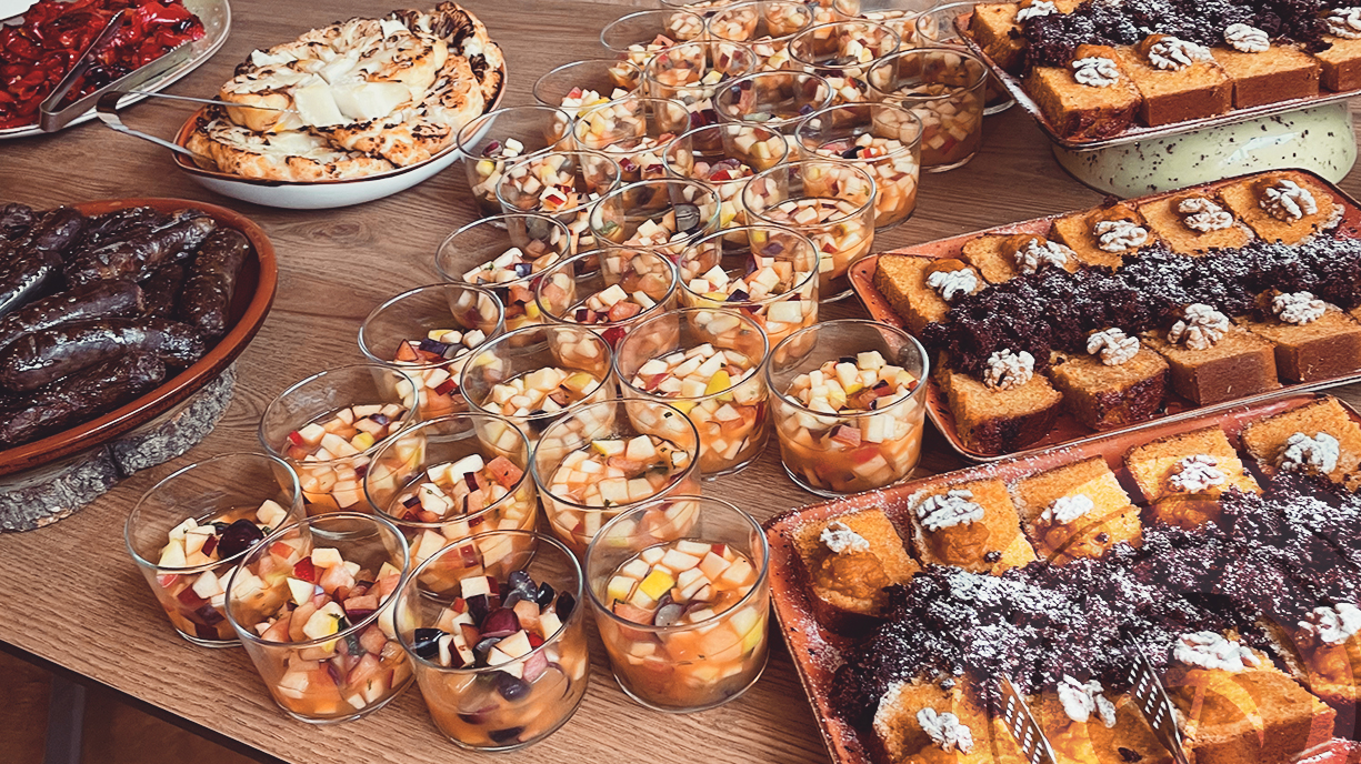 Buffet table with cups of fruit salad, trays of sponge cake topped with nuts and a dark crumble, a dish of sausages, and a toasted flatbread or pastry.