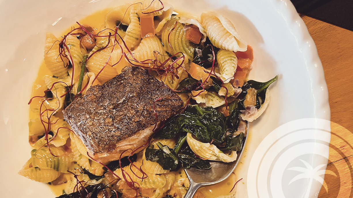 White plate with a rectangular, golden-browned piece in the centre, surrounded by short shell-shaped pasta, cooked green leaves and a light-coloured sauce, on a wooden table.
