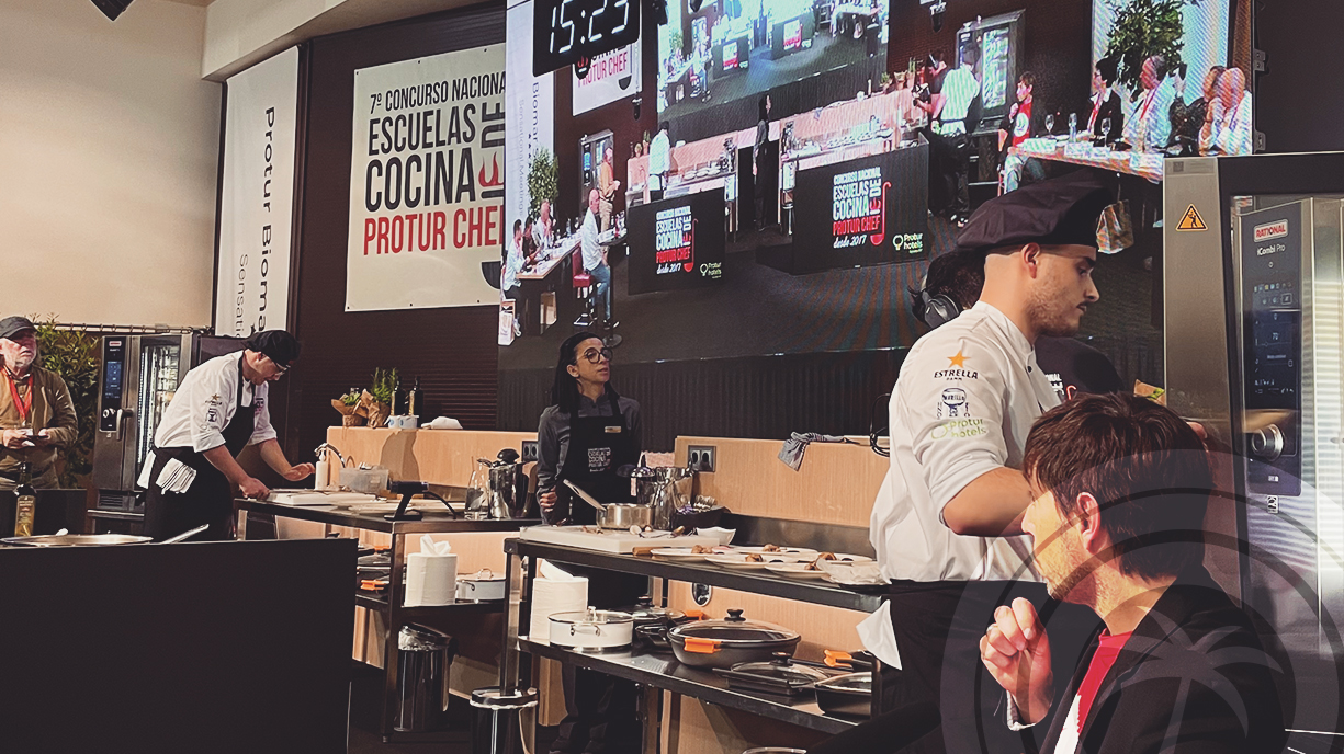 People in chef uniforms working at tables on a stage with a large screen behind