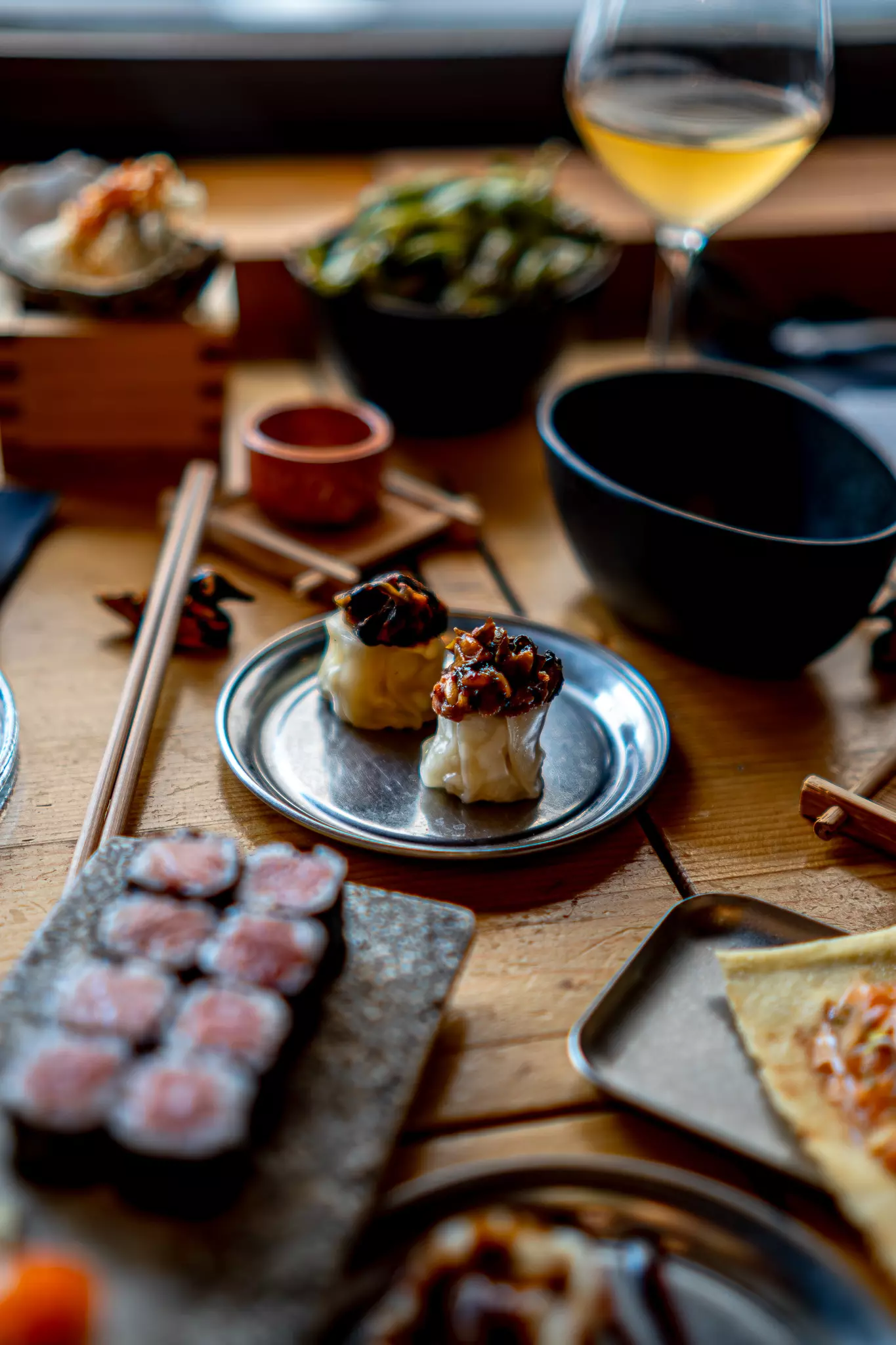 Sushi, dumplings and small dishes served on a table with chopsticks and a glass of white wine.